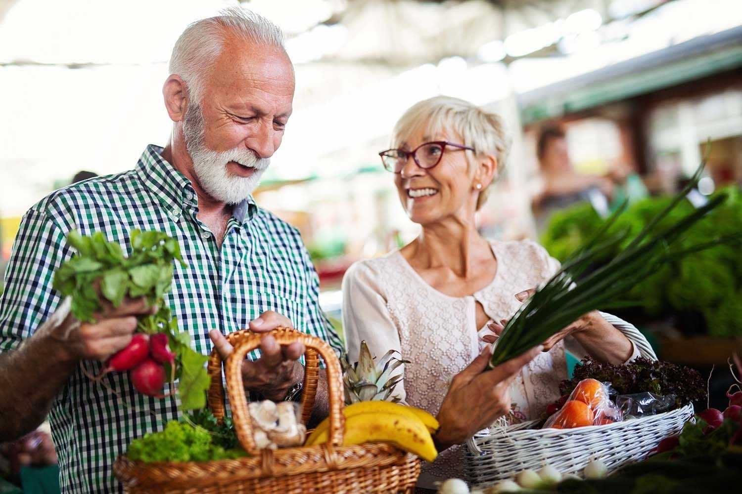 Auf dem Bild ist ein älteres Ehepaar zu sehen, das gerade Gemüse auf dem Markt kauft.